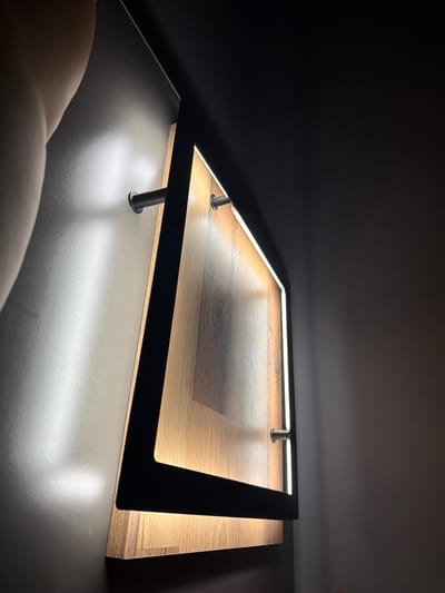 A low-angle, indoor shot shows a square, wooden surface with a dark brown, faux-wood-grain pattern. The surface is surrounded by a black frame with embedded LED lighting, supported by cylindrical metal posts. The LED lights are illuminated, casting a bright, warm light onto the wooden surface. The object sits on a flat, grey surface with a glossy sheen. The surrounding environment is dark, suggesting a dimly lit room or a closed space. No people are visible. There is no information to suggest the location is in Petarch, Bulgaria.