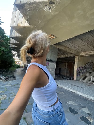 The image shows a blonde woman from the back, wearing a white crop top and blue jeans, standing in an abandoned, graffiti-covered building with concrete structures. She is looking up at the decaying architecture. The environment appears to be urban and neglected, with debris and overgrown vegetation visible. The specific location in Abaata, Abkhazia, is not discernible from the image alone.