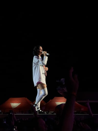 A medium shot captures a female singer on stage, holding a microphone to her lips and singing into it. She is wearing a long, white oversized shirt with "NOV" and "RO" visible in large letters on the front. Underneath the shirt, she has on a short pink garment, white fishnet stockings, and black platform shoes with ankle straps. Her dark hair is styled in a bob. The background is a dark, black stage with some red tents visible behind her, one with the word "AMBAR" in white lettering. In the foreground, the silhouettes of audience members with their hands raised are visible. The image was taken at the Auditorio Miguel Ríos in Rivas-Vaciamadrid, Spain.