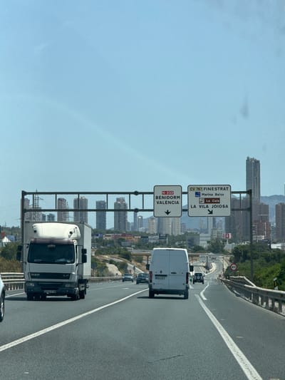 This is a photograph taken from a vehicle traveling on a highway, likely in Benidorm, Spain, based on the signage. The image is oriented vertically, with the road and surrounding scenery appearing upside down.

The primary subjects are two white vans and a white truck, all in motion on the highway. The truck is in the foreground, and its license plate reads "2032 FTW". The van behind it has a license plate reading "8864 NFG". In the distance, more vehicles are visible on the road, and tall buildings, characteristic of a cityscape, rise behind a line of trees and vegetation.

Several road signs are prominently displayed, providing directional information. One sign indicates "CV-767 FINESTRAT Marina Baixa La Cala LA VILA JOIOSA" with arrows pointing to the right. Another sign below it reads "N-332 BENIDORM VALÈNCIA" also with an arrow pointing to the right. A circular speed limit sign showing "80" is also visible.

The time of day appears to be daytime, with bright sunlight illuminating the scene and casting shadows. The sky is clear and blue, suggesting fair weather. The overall impression is that of a typical journey on a Spanish highway, with the urban landscape of Benidorm in the background and the road infrastructure taking center stage.