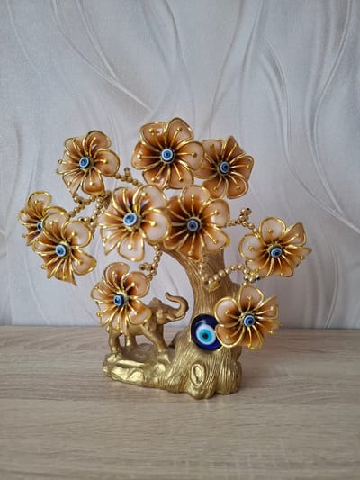 The image shows a decorative golden tree sculpture with translucent, petal-shaped ornaments featuring blue and white eye motifs. A small golden elephant figure is attached to the base of the tree. The sculpture is placed on a wooden surface against a textured, light-colored background. The scene appears to be an indoor setting, possibly a home, in Yablonovskiy, Russia, although there are no specific city identifiers present in the image itself.
