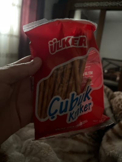 A red package of Ülker Çubuk Kraker, filled with pretzel sticks, is held in someone's hand. The package is slightly tilted, revealing the snack inside. The background includes a blurred view of furniture and a fuzzy white blanket. The brand name "Ülker" is prominently displayed in white lettering. "Çubuk Kraker" is written in a whimsical blue font.