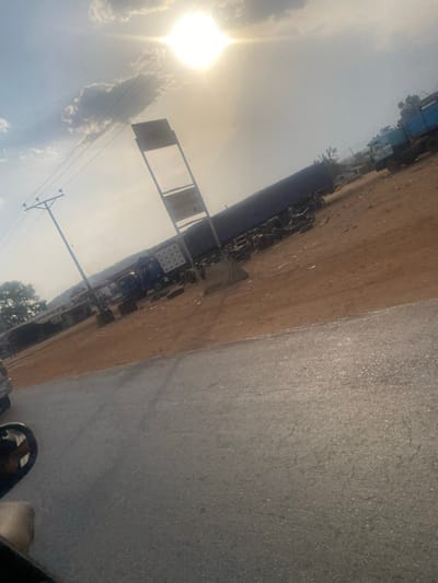 The image, viewed in its correct orientation, captures an outdoor scene along what appears to be a road or street, under bright daylight conditions. The perspective is slightly elevated, looking forward and to the left.

The sky, occupying the upper left portion of the frame, is bright and hazy with some scattered white clouds. A very intense, glowing sun is visible on the far left, casting a strong glare and creating a lens flare effect, indicating a clear, sunny day.

To the right, a segment of a paved road, likely asphalt, is visible. It is greyish in color with a textured surface and some minor cracks, suggesting typical road wear. The very bottom right corner shows a dark, rounded object, possibly part of a vehicle's tire or fender, indicating the photograph might have been taken from within a vehicle.

Running parallel to the paved road, in the center-left of the image, is a wide, unpaved strip of brownish-orange earth, appearing as a dusty roadside or an open field. This ground surface is uneven.

Along this dirt strip, in the midground, several objects and structures are present. A prominent feature is a tall, slender signpost with a rectangular metal frame holding two blank panels. On the lower panel, faint, partially legible text appears to read "SHREE" or a similar word. Behind the signpost, and extending further into the background, are numerous parked vehicles or pieces of equipment. Many appear to be two-wheeled vehicles (motorcycles or scooters) arranged in rows. There is also a distinct blue-colored structure, possibly part of a truck, container, or industrial machinery, visible among the other items.

On the far left side of the image, a utility pole, possibly made of wood or concrete, stands tall, with multiple power lines extending horizontally across the sky. In the distant background, a faint line of trees or foliage borders the horizon.

The scene is static, with no explicit activity taking place, conveying a sense of a quiet, possibly semi-urban or industrial roadside area bathed in strong sunlight.
