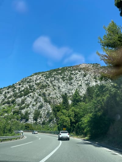 A wide, curving asphalt road, with white lane markings, passes through a mountainous, green landscape under a bright blue sky. The road is bordered by a metal guardrail on the left and dense green trees and vegetation on the right. Several cars are visible on the road, including a white SUV in the foreground on the right lane, a silver sedan and a white hatchback further ahead on the left lane, and a white sedan in the distance. The mountainside on the left is covered with gray rocks and scattered green vegetation. The sky is clear with a few wispy clouds. The overall scene suggests a scenic drive through a natural environment.