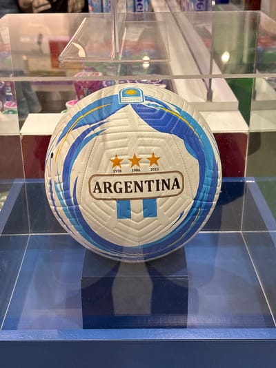 The image shows a close-up of a white soccer ball with blue and yellow accents, displayed in a glass case. The ball has the word "ARGENTINA" printed on it in a golden oval emblem, along with three orange stars and the years "1978", "1986", and "2022". The ball is positioned on a dark blue rectangular pedestal, which is inside a larger, clear glass display case. The background is blurred, but appears to be an indoor setting with shelves and possibly other merchandise. There is no discernible connection to the city of Ezeiza, Argentina.