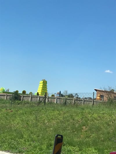 A vibrant green grassy field stretches under a clear blue sky. A wooden fence runs across the field, partially obscured by the grass. A tall, multi-tiered yellow structure, potentially a decorative tower, rises from behind the fence. A building with a tan facade is visible in the background. A small white cloud floats in the sky. A sign with yellow stripes is partly visible in the lower right.