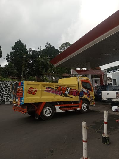 The image depicts an outdoor scene at a gas station in Ariyalai East, Sri Lanka, under an overcast sky. The primary subject is a yellow dump truck, positioned on a dark asphalt surface, facing right. The truck features an orange chassis, white multi-spoke wheel rims, and extensive graphic decals. The side of the dump bed displays a red "02" and large stylized text reading "BERAS ANUOKA". The cabin door contains graphics including "It's My 4life" and other lettering. The rear section of the dump bed on the left side shows a large rectangular graphic featuring a muscular man and text in Indonesian.
In the background to the left, a dark stone wall with a black and white pattern on its upper section is visible, with dense green foliage and tall trees behind it. To the right, a gas station canopy with a red top edge and a grey underside is supported by grey pillars. Signs under the canopy display "PERTALITE" and "PERTAMINA". Partially visible under the canopy are a dark blue SUV or pickup truck and a white pickup truck, both facing away from the camera. A white object, possibly a motorcycle helmet, rests on the ground near the white pickup truck. In the lower right foreground, two white bollards with red stripes are visible, connected by a rope. No individuals are present in the scene, and all vehicles appear stationary.