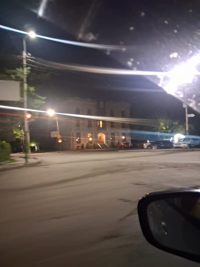 The image captures a nighttime view of a street in Yeghegnadzor, Armenia, from the perspective of a vehicle. The foreground is dominated by a car's side mirror, reflecting a blurry interior and the street ahead. The street itself appears wet, with faint reflections of streetlights.
In the mid-ground, a building with illuminated windows stands prominently. It has a classical architectural style with arches and columns. Several cars are parked along the street in front of the building, including a red car, a dark SUV, and a white van. A traffic light is visible on a pole, appearing to be red. Streetlights illuminate the scene, casting long streaks of light due to motion blur. Trees and foliage are present on the left side of the street. The sky is dark, suggesting it is late at night.