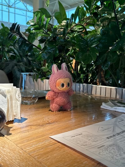A pink, plush bunny doll with large, round eyes and a smiling expression stands on a wooden table in what appears to be a restaurant. The doll is positioned in the center of the frame, facing forward. To the left of the doll, there is a clear glass with water and a stack of white napkins. To the right, a white napkin is folded with a fork and knife placed on top. In the background, lush green plants with broad leaves are visible, creating a naturalistic ambiance. The table surface shows a natural wood grain pattern, and the overall lighting suggests an indoor setting, possibly illuminated by natural light from above through a glass ceiling, as hinted by the blue-tinted light in the upper left corner. A menu with illustrations and text is laid out on the table in the foreground. The setting is likely the Agalarov Estate in Russia, a resort area known for its restaurants and leisure facilities.