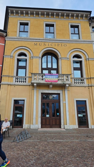 The image shows the facade of a yellow municipal building, likely the town hall, in Riva del Garda, Italy. The building has a classical architectural style with arched windows and decorative stonework. A small rainbow-colored flag with the word "PACE" (Italian for peace) written on it in colorful letters is displayed on a balcony. The flag appears to be a symbol of peace or LGBTQ+ pride. The building's signage reads "MUNICIPIO," which means municipality.

In the foreground, a person is sitting on a chair outside the building, and another person's legs and feet are visible on the right. A cobblestone street or plaza is adjacent to the building, and a bicycle rack is also present. The sky is visible in the upper left portion of the image, suggesting it is daytime. The overall impression is a peaceful, everyday scene in an Italian town.