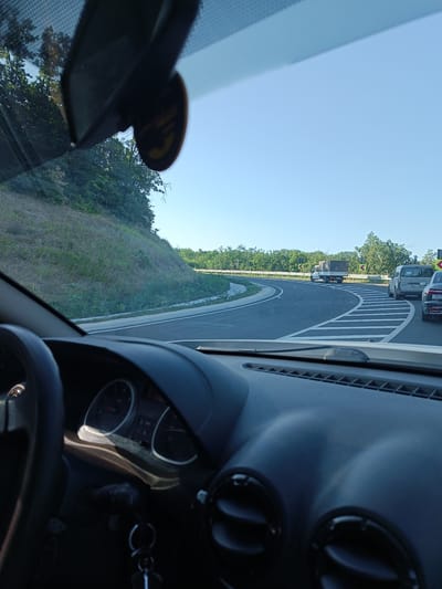The image depicts a view from the interior of a car driving on a road near Rusca, Moldova. The driver's side dashboard and steering wheel are visible in the foreground. The road ahead curves to the right, with lane markings indicating a traffic flow. Several vehicles are present on the road, including a light-colored truck carrying a large, dark rectangular object, a white van, and a dark sedan. The landscape surrounding the road consists of a grassy embankment on the left and trees on the right. The sky is clear and blue, indicating daytime.