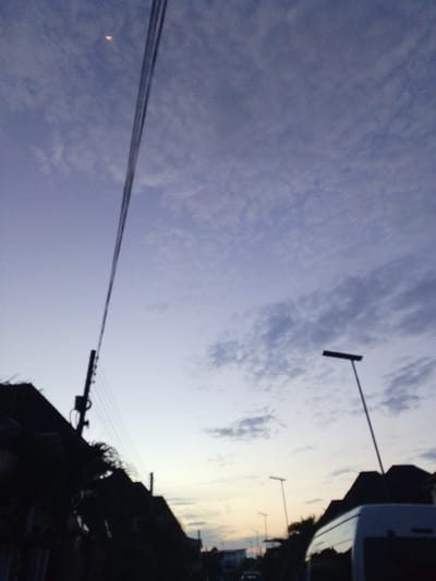 The image captures a twilight scene in Rumuomasi, Nigeria, with a dusky purple sky populated by wispy clouds. A sliver of the moon is visible in the upper left quadrant. A dark silhouette of utility poles with suspended wires cuts diagonally across the frame from the left, disappearing upwards. In the foreground, silhouetted buildings with pointed roofs and dense foliage are visible on the left side. A white van, partially visible, is parked on the right side of a road. Further in the background, streetlights are intermittently illuminated. The overall impression is one of a quiet, transitional moment at dusk in an urban or suburban setting.