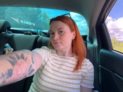 A woman with red hair is sitting in the back seat of a car, taking a selfie. She is wearing a white and yellow striped shirt. Her left arm, which is visible in the foreground, has a large floral tattoo. There are several small toy vehicles on the seat behind her. Through the rear windshield, trees and a bright blue sky with white clouds are visible. The location is the city of Забегалово, Russia.