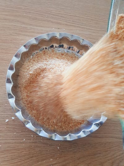 The image shows a close-up, top-down view of a fluted metal pie dish filled with breadcrumbs. A blurry stream of orange-colored breadcrumbs is being poured from a clear glass container into the dish, obscuring a portion of the dish and its contents. The dish is placed on a wooden surface, likely a table, with scattered white crumbs visible on the wood. The scene is set indoors, suggesting a kitchen environment, but no specific location in Kheyrabad, Iran, can be identified from the visual information provided. The lighting appears to be natural or overhead artificial light, casting subtle reflections on the metal dish.
