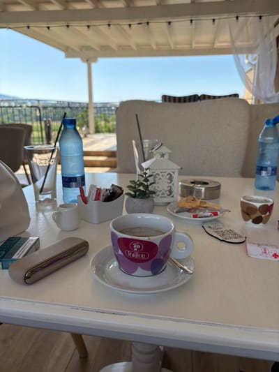 This is a photograph of a table with various items on it, set outdoors under a canopy. The setting appears to be a cafe or restaurant in Ognyanovo, Bulgaria, given the outdoor seating and the view of greenery and distant hills.
On the table, from left to right, is a pack of cigarettes, a silver and beige electronic cigarette device, a white sugar dispenser filled with sachets of sugar and sweeteners, a small white pitcher, a glass with a straw, a blue bottle of water, a small potted plant, a white decorative lantern, an ashtray with some remnants, a saucer with a spoon and a candy, a coffee cup with a spoon on the saucer, and a smaller cup with a brown and beige pattern, a QR code coaster, and a packet of sugar or sweetener. The coffee cup has a purple and pink patterned design with the word "Portion" visible.
The background shows a blurred view of outdoor seating with cushioned furniture and white curtains, suggesting a relaxed, open-air environment. The sky is clear and blue. The overall impression is one of a leisurely setting, possibly for coffee or drinks.