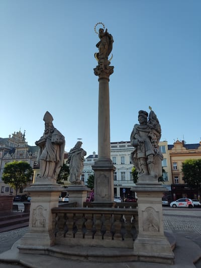 The image depicts a stone monument in Kolín, Czechia, featuring multiple statues. The central element is a tall column topped with a statue of a woman with a halo, possibly the Virgin Mary. At the base of the column, on either side, stand two large statues of male figures. One on the left appears to be a bishop, holding a miniature building and a book. The statue on the right depicts a warrior-like figure holding a flag. The monument is surrounded by a balustrade and is situated in a public square or plaza. In the background, several buildings are visible, including a building with ornate architecture and other structures with multiple stories and windows. Trees are present, and parked cars are visible on the street behind the monument. The sky is clear and blue. The overall scene suggests a historical or religious monument in an urban setting during daylight hours.