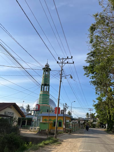 The image captures a street scene in Alurcucur, Indonesia, under a bright blue sky with wispy clouds. Overhead, a dense network of power lines crisscrosses the frame, supported by utility poles. To the left, a mosque with a distinctive green and white striped minaret and a large dome is visible. Adjacent to the mosque are several colorful buildings, some with painted facades. A water tank with "PERTAMINA PEDULI" branding stands on a metal structure near the mosque. A dirt road runs alongside the buildings, leading to an asphalt road in the foreground. A person is seen riding a motorcycle on the asphalt road, heading away from the camera. Lush green trees line the right side of the road, casting shadows on the scene. The overall impression is of a sunlit, modest urban or semi-urban environment.