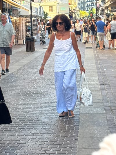 The image shows a street scene in Chania, Greece, likely within the Old Town, characterized by its cobblestone pavement. In the foreground, a woman with dark, curly hair and sunglasses walks towards the viewer, wearing a white sleeveless top with a polka dot pattern and loose-fitting light blue pants. She is carrying a light-colored handbag with leather accents in her right hand. Her feet are adorned with dark sandals, one of which reveals a tattoo on her foot. Behind her, other people are visible, some walking in the same direction, others strolling in the opposite direction. A man in a light-colored short-sleeved shirt and dark shorts is walking away from the camera to the left. Shops line the street, displaying merchandise such as sunglasses. The overall atmosphere suggests a busy pedestrian area on a sunny day.