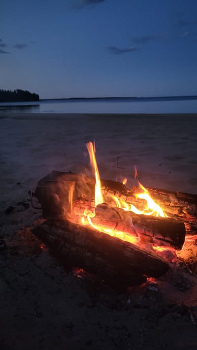 A campfire burns on a sandy beach at dusk, with the ocean and a distant treeline visible in the background. The sky is a deep blue, fading to a lighter hue near the horizon. The fire illuminates several logs and embers, casting a warm glow on the surrounding sand. A metal cup sits next to the fire.