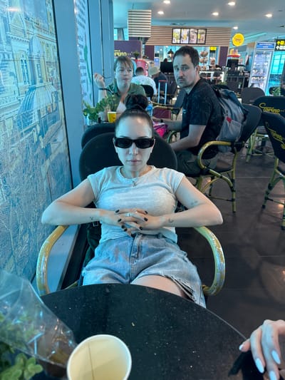 The image depicts a cafe or waiting area at Cairo International Airport, Cairo, Egypt. In the foreground, a woman with sunglasses, a gray t-shirt, and denim shorts is seated. She has black nail polish and tattoos on her arm. A black table is nearby, with a disposable cup and plastic bag. Behind her, a man wearing a patterned shirt and a backpack is seated. Further back, another woman in a green shirt and a man are visible. The background shows airport signage and other commercial establishments. The floor has a graphic design resembling a cityscape.