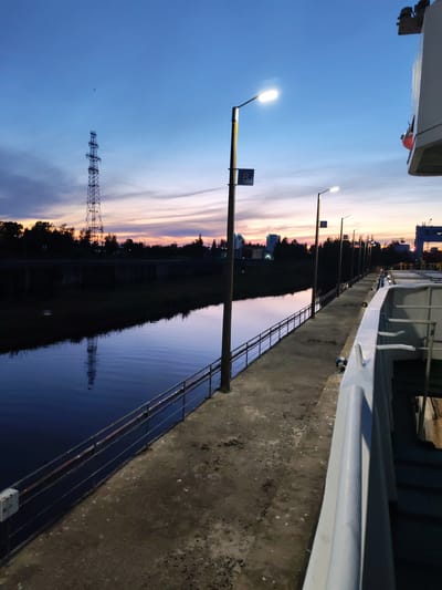 The image depicts a waterfront area at dusk, likely in the city of Sheksna, Russia, given the context provided. On the left side, a body of dark water reflects the twilight sky and a tall radio tower in the distance. A concrete walkway runs along the water's edge, separated by a metal railing. Several lampposts line the walkway, casting a warm glow on the scene. On the right side, the edge of a large vessel, possibly a ship or barge, is visible, with its white railing and some metal structures. The sky is a gradient of deep blue at the top, transitioning to hues of orange and pink near the horizon, indicating the sun has recently set. Silhouetted trees and distant buildings are visible on the far bank of the water. The overall atmosphere is calm and serene.