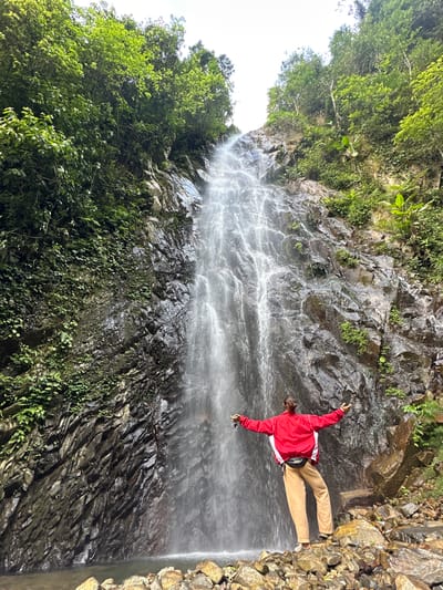 A lone individual stands facing a tall, cascading waterfall in Tu Le, Vietnam. The person, viewed from behind, wears a red and white long-sleeved jacket, light brown pants, and a black fanny pack, with arms outstretched to the sides, holding sunglasses in the left hand. They are positioned on a collection of angular brown and grey rocks at the base of the waterfall. The powerful waterfall descends a steep, dark, and wet rocky cliff face, forming a shallow pool at its foot. The surrounding slopes and the top of the cliff are densely covered with lush, vibrant green vegetation, including various trees and smaller plants. The sky above is bright and appears overcast.