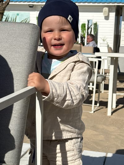 A vibrant outdoor portrait captures a young child, likely a toddler, positioned in the foreground, smiling broadly at the camera. The child wears a dark blue beanie featuring a small white square emblem with a stylized black letter, possibly 'B' or 'R'. They are dressed in a light-colored, possibly beige or khaki, long-sleeved shirt and matching trousers. The child's left hand is seen gripping the white frame of an outdoor chair or table, a portion of which is upholstered in a light grey fabric in the lower left.
The setting appears to be a sunny outdoor area, possibly a cafe or restaurant terrace in Middelkerke, Belgium. The ground beneath the furniture is sandy, hinting at a beachside or coastal location. In the mid-ground, an adult male is partially visible and out of focus, seated among white outdoor tables and chairs. Other white structures, possibly beach cabins or resort buildings, are discernible in the background.
Strong, sharp shadows cast by the furniture indicate bright sunshine and clear weather, suggesting the time of day is likely midday or early afternoon. The child's wide smile conveys a joyful and relaxed mood.