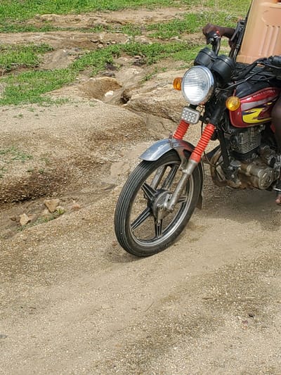 The image shows a close-up, angled view of the front of a motorcycle parked on a dirt and gravel path. The motorcycle has a red and yellow fuel tank, a chrome headlight, and orange turn signals. Red accordion-style covers are visible on the front suspension. A person's arm and hand, wearing a dark sleeve, are resting on the handlebars, suggesting they are either about to ride or have just dismounted.

To the left of the motorcycle, there is a patch of green grass and some rough, uneven terrain, possibly indicating a roadside or an unpaved area. The ground where the motorcycle is parked is composed of small, light-colored pebbles and dirt, with some darker patches suggesting dampness or oil stains.

The scene appears to be outdoors, likely in a rural or semi-rural area given the unpaved ground and vegetation. There are no clear indicators of the time of day or specific weather conditions, although the lighting suggests it is daytime. The overall impression is a casual, everyday scene in a location like Kabwir, Nigeria. No text is visible in the image.