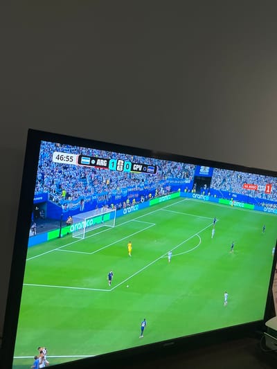 The image captures a television screen displaying a soccer match. The game appears to be Argentina vs. CPU, with the score currently 1-0 in favor of Argentina at 46:55 into the match. The stadium is filled with spectators wearing blue and white, likely supporting Argentina. The field is green, and there are several players in various positions on the field. The TV is placed on a wooden surface, possibly a table or stand, in a dimly lit room. The background is a plain, dark gray wall. A small portion of a white and green object is visible on the right side of the image, potentially a notebook or a box, and a green cord extends from it. The scene is domestic, as indicated by the presence of a television in a room. There is no visible indication that this scene is located in the city of Debelets, Bulgaria; this information is not derivable from the provided image.