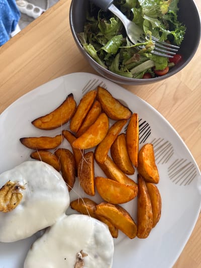 This media file shows a close-up, overhead view of a meal set on a wooden table. The meal consists of two main components: a bowl of salad and a white plate filled with potato wedges and melted cheese.

The salad, in a dark grey bowl, appears to be a mix of leafy greens and cherry tomatoes. A silver fork rests within the salad.

The white plate, which has a decorative pattern of grey lines, holds a generous portion of golden-brown potato wedges. Some of the wedges show signs of seasoning, possibly paprika. Towards the right side of the plate are two circular portions of melted cheese, with some of the cheese stretched between the wedges and the cheese portions. There are also a few charred pieces visible within the cheese.

The scene suggests a casual meal, likely lunch or dinner. The lighting is natural, indicating it might be daytime. There are no visible people, text, or specific location cues within the image itself, other than the implied setting of a table. However, based on the provided location context of Almada, Portugal, it can be inferred that the meal is being enjoyed there.
