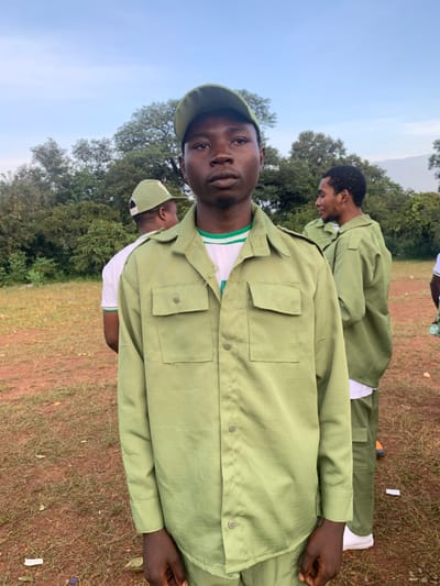 The image captures an outdoor scene within the city of Kubi A, Nigeria, featuring an open field environment. In the foreground, a young adult male of African descent, estimated to be between 20 and 30 years old, stands facing the viewer with a neutral expression. He is dressed in a light olive-green long-sleeved uniform shirt featuring two chest pockets and shoulder epaulets, paired with a matching baseball cap. A white t-shirt with green and white striped trim is visible underneath the uniform shirt.
In the mid-ground, to the subject's left, another male is partially visible from behind, wearing a white t-shirt and a similar olive-green cap. To the subject's right, a third male stands facing right, wearing a full light olive-green uniform, including a long-sleeved shirt, trousers, a white undershirt, and white athletic shoes. A fourth individual is partially visible at the far right edge. The uniforms worn by the individuals are consistent with those utilized by participants in Nigeria's National Youth Service Corps (NYSC) program.
The ground beneath the subjects is a reddish-brown dirt surface, interspersed with patches of sparse green grass and small pieces of white debris. The background is dominated by a dense line of various green trees and foliage, situated beneath a clear, light-blue sky, indicating a daytime setting.