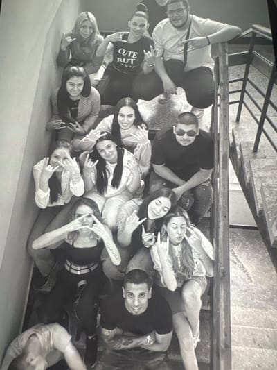 A black and white image shows a group of approximately 16 people lying closely together. They appear to be posing for a photograph, as many are looking directly at the camera, smiling, and making hand gestures such as the "rock on" sign. Some individuals are wearing sunglasses, and a person in the middle wears a shirt that says "CUTE BUT." Above them, there is a metal railing and a portion of what seems to be a concrete staircase. The image is taken in the city of Petarch, Bulgaria.