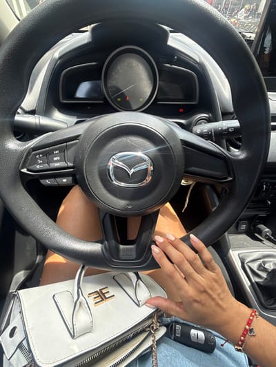 The image depicts the interior of a Mazda vehicle from the driver's perspective. A person with tan skin is seated, with their right hand resting on a white structured handbag, which has a gold "EE" or "E3" logo on its front flap. The hand displays manicured nails with a light pink, iridescent finish, and a red beaded bracelet with a black clover charm is worn on the wrist. A black car key fob is visible near the handbag on the person's lap. The black steering wheel, bearing the Mazda logo and an "SRS AIRBAG" inscription, dominates the foreground, featuring multimedia controls on its left spoke. The instrument cluster behind the steering wheel shows an analog speedometer at zero, with illuminated green and red indicator lights. Portions of the center console, including a gear selector area and a USB port, are visible to the right. A blurred, sunlit outdoor environment is visible through the windshield. The scene is situated in Pitalito, Colombia.