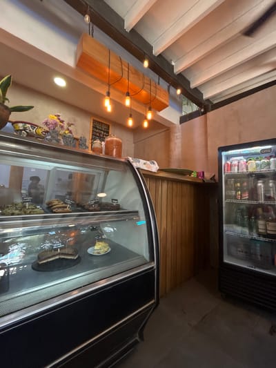 The image shows the interior of a bakery located at latitude: 9.629, longitude: -85.152 in the Cobano District of Costa Rica. The scene features a glass display case containing pastries and cakes, including a large chocolate cake, and a smaller slice of pie. The display case is positioned in front of a wooden wall, which is adjacent to a refrigerator filled with various drinks, including bottled beers and other beverages. The ceiling is lined with white wooden panels, illuminated by a string of hanging lightbulbs. The bakery appears to be empty of customers.