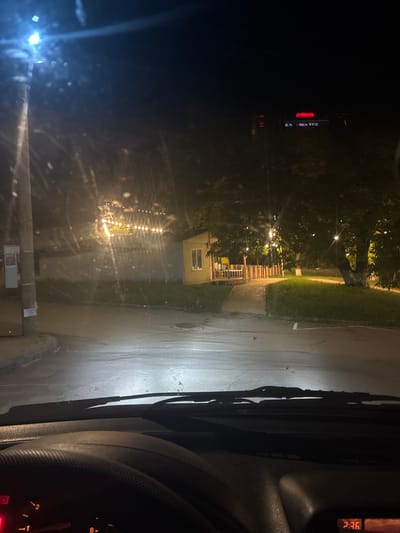 The image, taken at night from inside a vehicle, shows a view through the windshield. In the foreground, the dashboard and windshield wipers are visible. The scene outside depicts a street with a lamppost on the left and a small building with a colorful fence in the middle ground. String lights are illuminated above the building. Trees are present in the background. A digital display on the right side of the dashboard shows "2:36". The location is identified as Gorna Malina, Bulgaria.