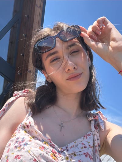 The image shows a young woman taking a selfie outdoors. She is wearing a floral dress and a necklace with a cross pendant. She is holding her sunglasses above her head with one hand. The background consists of a blue sky and a wooden structure. The image does not contain any information about the city of Sharovechka, Ukraine.