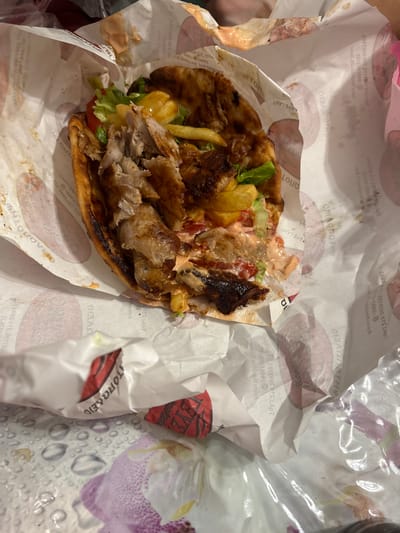 The image shows a close-up, overhead view of a partially eaten gyros wrap resting on branded paper. The wrap is filled with grilled meat, french fries, lettuce, and tomatoes, and appears to be drizzled with a creamy sauce. The paper wrapper has Greek text and logos, including "ΑΤΟΠΩΛΕΙΟ" and "ΓΕΥΣ", indicating it is from a food establishment in Greece. The lighting suggests it is daytime, and the setting appears to be casual, possibly a meal at home or on the go. There are no people visible in the image, but a portion of a hand with pink fingernails is visible on the left edge. The overall impression is a somewhat messy, delicious-looking meal.