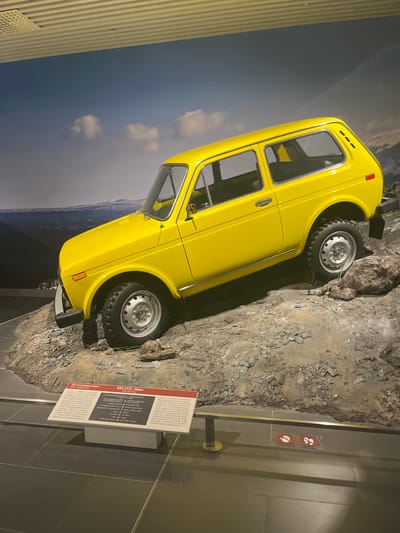 A bright yellow Lada Niva is displayed on a simulated rocky terrain in a museum setting in Verkhnyaya Pyshma, Russia. The car is positioned at an angle, with its front wheels slightly elevated on the rocks, suggesting off-road capability. A factual plaque is placed in front of the display, providing information about the vehicle. The background features a large photographic mural of a cloudy sky and mountainous landscape. The floor in front of the display is tiled. Two red circular prohibition signs, one showing hands and the other feet, are visible on the floor near the front of the exhibit. The scene appears to be indoors, possibly within the "Battle Glory of the Urals" museum complex, which houses a significant collection of Soviet-era military and civilian vehicles. There are no people visible in the frame.