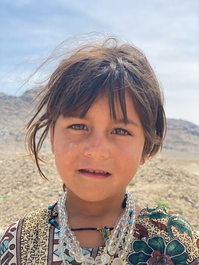 A close-up shot captures a young girl, likely of Afghan descent, in an outdoor setting near Ashoqah, Afghanistan. Her slightly dishevelled brown hair frames her face, and her eyes are hazel. A light smudge of dirt or powder is visible on her left cheek. She wears a patterned tunic or dress in beige, blue, and green hues, along with multiple strands of clear and blue beaded necklaces. In the background, a dry, mountainous landscape under a bright sky is visible.