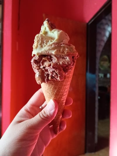 A hand holds a waffle cone filled with two scoops of ice cream in front of a red wall. The top scoop appears to be pistachio while the lower scoop is a dark chocolate. The hand is positioned to the left of the frame and a dark doorway, with a blurred background, is to the right. The image was taken in El Talar, Argentina, at the coordinates -34.473, -58.639.