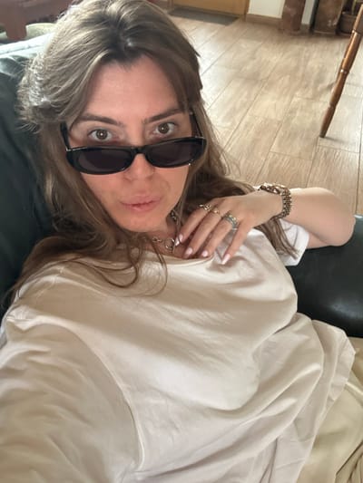 An adult female is depicted indoors, likely in Sighnaghi, Georgia, reclined on what appears to be a dark-colored sofa or armchair. She is looking directly at the camera with her lips slightly parted. She has light brown hair styled with bangs and is wearing dark, rectangular sunglasses positioned mid-nose, revealing her eyes above the frames. Her attire consists of a light, possibly off-white or beige, short-sleeved t-shirt. Her left hand is raised, showcasing multiple rings on her thumb, index, middle, and ring fingers, and a silver-toned chain necklace with a circular clasp is visible around her neck. A gold-toned wristwatch with a metal link band is present on her right wrist. The background features light wooden floorboards, a partial view of a wooden chair leg, and a light-colored wall with a wooden door frame.