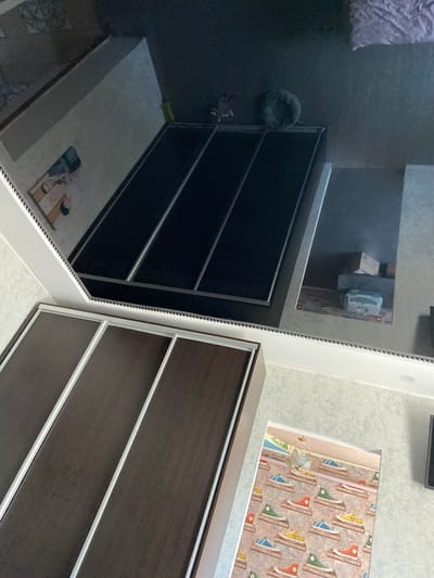 The image is a close-up, reflected view of a room, likely a bedroom or dressing area, as evidenced by the presence of a wardrobe and the overall interior decor. The primary focus is on a dark, reflective surface, which appears to be the mirrored door of a wardrobe. This surface reflects various elements of the room.
In the reflection, we can see what looks like a bed or a large, dark rectangular surface, possibly a platform bed with a dark mattress, positioned in the upper portion of the mirrored surface. To the left of this, a framed picture or artwork featuring two individuals is visible. Further to the right, a light-colored, rounded object, possibly a pet bed or a stuffed animal, is present. Behind that, on what appears to be a shelf or surface, are several boxes, one with a white, perforated appliance that could be a humidifier or air purifier.
Below the reflected scene, in the foreground, is the actual wardrobe, with its dark wood-effect doors and silver trim. The wardrobe has sliding doors, and we see at least three panels. The texture of the wallpaper on the wall to the right of the wardrobe is also visible, featuring a repeating pattern of colorful sneakers.
The image is taken from a slightly high angle, looking down and into the reflection. The lighting is somewhat dim, creating a moody atmosphere. The exact location in Rozkvit, Ukraine, cannot be determined from the visual information provided in the image.
