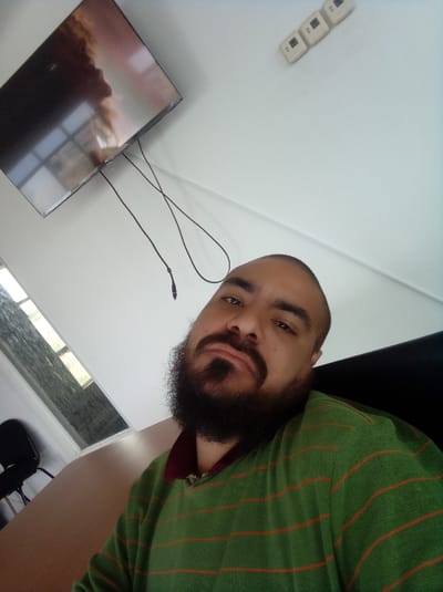 A close-up selfie of a person with a dark beard and mustache, wearing a green and orange striped sweater with a maroon collar. The person is indoors. Behind the person is a white wall with a flat-screen television mounted on it. Three white electrical outlets are also visible on the wall. A window is to the left with a black chair visible, the city and town information is indeterminable, the country of Venezuela is accurate based on the model but there is no city to identify it.