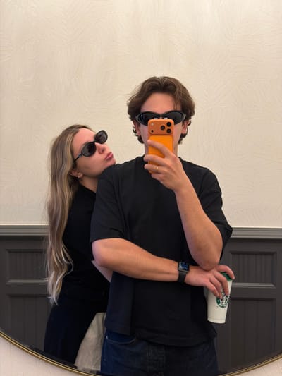 A young person, wearing sunglasses and a black t-shirt, holds an orange smartphone in their right hand and a Starbucks cup in their left. They are taking a selfie in a large, oval mirror. The mirror reflects another person, also wearing sunglasses and a black t-shirt. The background is a wall with a subtle leaf-patterned wallpaper and a dark-colored door. The lighting suggests it is daytime. The people appear relaxed and casual. The Starbucks cup is white with the brand's logo visible. The person taking the selfie has a ring on their right ring finger. A smartwatch is visible on their left wrist. The scene is likely indoors, possibly in a hotel room or apartment.