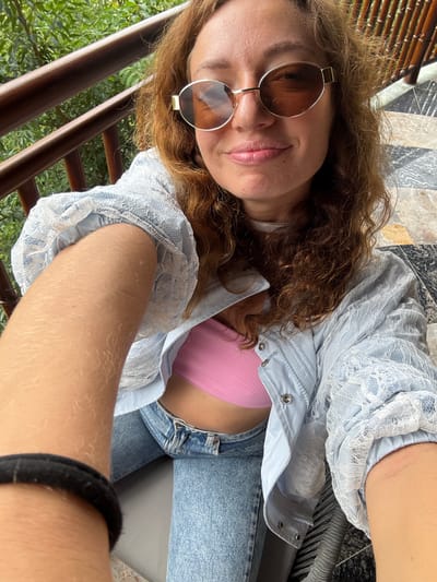 A woman is captured in a close-up, high-angle selfie. She has light brown, curly hair and wears brown-tinted sunglasses with gold-toned frames. Her lips are slightly parted in a faint smile. She is dressed in a light blue jacket with white lace detailing on the sleeves and shoulders, worn over a pink ribbed crop top, and blue denim jeans. Her left arm, positioned in the foreground and extending towards the camera, shows fair skin with visible light hair, and a black fabric band is present on her wrist. The background features a wooden railing on the left, beyond which is lush green foliage. On the right, a structure of vertical wooden slats is visible, adjacent to a patterned tiled floor in shades of grey and black. The scene depicts an outdoor setting, located in Púng Luông, Vietnam.