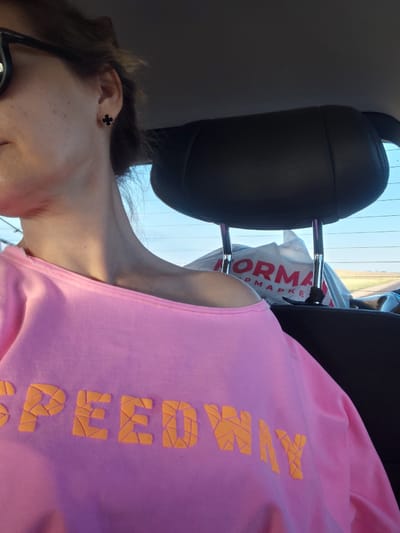 The image shows a close-up of a person sitting in the passenger seat of a car. The person is wearing a pink off-the-shoulder sweatshirt with the word "SPEEDWAY" printed in orange lettering. The sweatshirt has a textured, almost mosaic-like design on the letters. The person is also wearing black sunglasses. In the background, a black leather headrest is visible, and behind that, a shopping bag with the word "NORM" printed on it can be seen. The scene appears to be outdoors, as suggested by the sky visible through the car window, which shows a clear blue sky and some distant fields. The exact location within the city of Молодёжный, Kazakhstan, cannot be determined from the image alone.