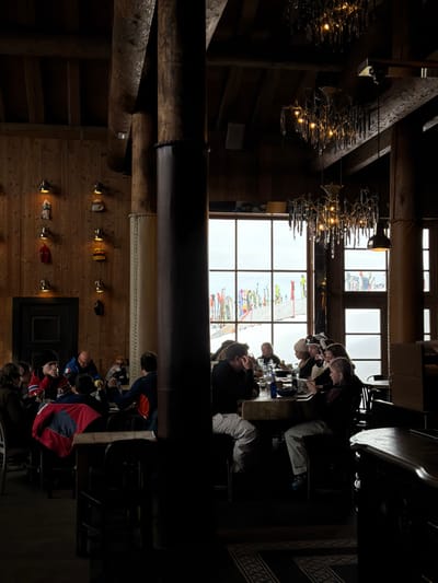 The image displays the interior of a ski lodge or restaurant in Les Allues, France. The space features dark wooden plank walls and ceiling beams, with several thick, dark wooden support columns, some wrapped in a padded material. Ornate chandeliers with multiple light sources hang from the ceiling, alongside smaller wall-mounted sconces illuminating decorative items, possibly hats or mittens, displayed on hooks along the left wall. A dark wooden door is also present on the left.
Approximately 15-20 individuals are seated at several rectangular wooden tables throughout the room. They are dressed in winter clothing, including jackets, sweaters, and some wearing beanies or ski hats. Actions include dining, drinking from glasses and bottles, and social interaction, with individuals looking at each other or at items on the table, and one person on the right viewing a smartphone.
Large, multi-paned windows dominate the background, revealing a snow-covered outdoor landscape under an overcast sky. Beyond the windows, numerous colorful skis and snowboards are vertically planted in the snow, indicative of a ski slope or equipment staging area. The floor is dark, with a patterned rug visible beneath tables on the right.