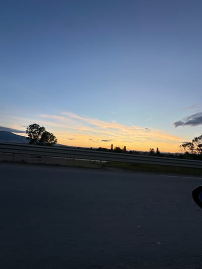 A wide shot shows a sunset over Kazichene, Bulgaria, with a highway in the foreground. The sky transitions from a clear, pale blue at the top to streaks of orange and yellow near the horizon, indicating dusk. A mountain range is visible in the far left distance. Silhouettes of trees and a line of smaller trees with some buildings behind them are spread across the middle ground. A metal guardrail runs horizontally through the scene, separating the highway from the landscape. The highway surface in the foreground is dark asphalt, suggesting it is either evening or the lighting is dim. The image appears to be taken from a moving vehicle, as suggested by the slightly blurred foreground and the edge of a side mirror visible in the bottom right corner.