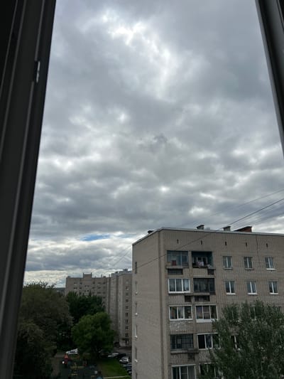 This is a vertical, wide-angle shot taken from what appears to be a window, looking upwards and outwards towards a cloudy sky and residential buildings. The perspective is tilted, giving a sense of looking down from a height.

The primary subjects are two multi-story brick apartment buildings, viewed from the side and from behind. The buildings have numerous windows, some of which appear to be dark or empty, while others show signs of occupancy with various items visible through them. Some windows have exterior air conditioning units. A large tree with dark green foliage partially obscures the right side of the foreground building. In the distance, more apartment buildings are visible, interspersed with trees. A glimpse of a playground with colorful equipment and some parked cars can be seen between the buildings and trees in the lower right portion of the image.

The scene is set outdoors in a residential area. The sky is overcast with dense, gray clouds, suggesting it is daytime, likely afternoon, with the sun not shining brightly. There is a small patch of blue sky visible to the right, breaking through the clouds. The overall mood is somewhat subdued due to the cloudy weather.

There is no discernible text in the image. The focus is on the architecture and the atmospheric conditions.