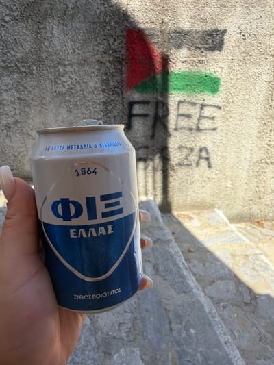 A hand with manicured fingernails holds a white and blue can of "FI FIELLAS" beer in front of a rough, gray concrete wall with graffiti. The graffiti depicts the Palestinian flag colors (red, black, and green) and the words "FREE GAZA" spray-painted in black. The background shows a stone staircase with sunlight casting shadows. The scene appears to be an outdoor urban environment.